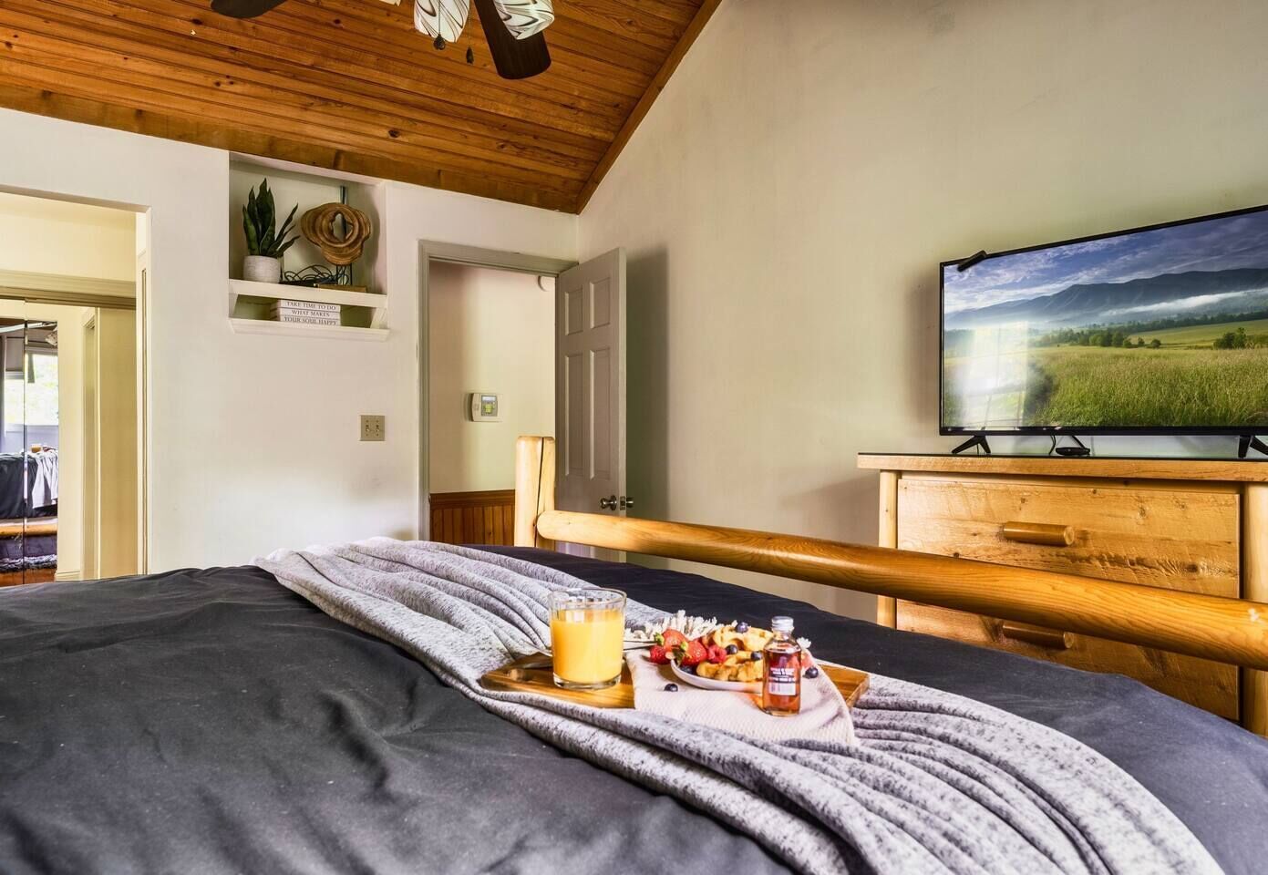 Een slaapkamer met een bed, een houten hoofdbord, een televisie, een plank met decoratieve objecten, en een deur naar een andere kamer.