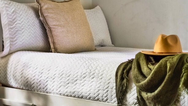 Una cama con cubrecama blanco acolchado, almohada de arpillera y manta verde.