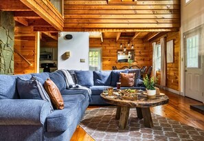 A cozy cabin interior with a blue sectional sofa, a round wooden coffee table with snacks, and a stone wall. - Rustic Cabin with Fire Pit and Hot Tub (Pigeon Forge)