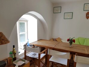 Ein Essbereich mit Holztisch, weißen Stühlen und einer grünen Couch. Ein Fenster bietet Ausblick, links ist eine Lampe zu sehen.
