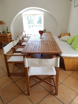 A dining area with a wooden table and chairs, a vase with red flowers, and a bed with green pillows.
