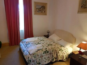 Ein Schlafzimmer mit einem Bett, einem Nachttisch, einer Lampe, einem Fenster mit Vorhängen und einem Bild an der Wand.