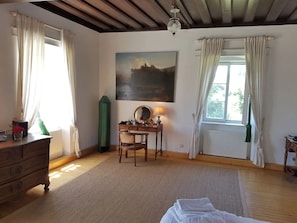 A room with a wooden dresser, a desk with a mirror, a painting on the wall, and a window with curtains.