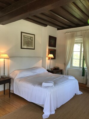 Ein ordentlich bezogenes Bett mit weißen Laken, ein Nachttisch mit Lampe und ein Fenster mit Vorhängen.
