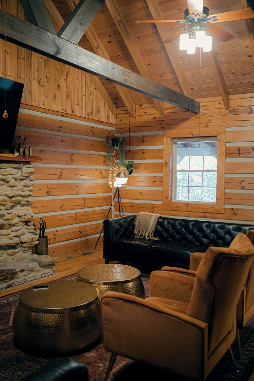A log cabin interior with a black leather sofa, a round woven coffee table, and a stone wall.