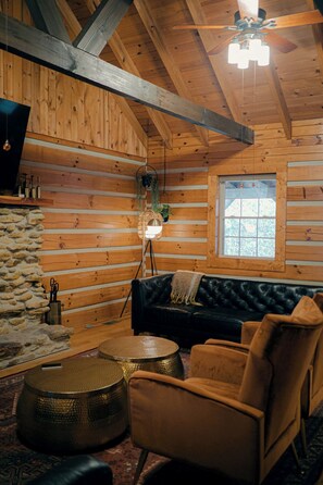 Interior d'una cabana de fusta amb un sofà de cuir negre, una taula de centre rodona teixida i una paret de pedra.