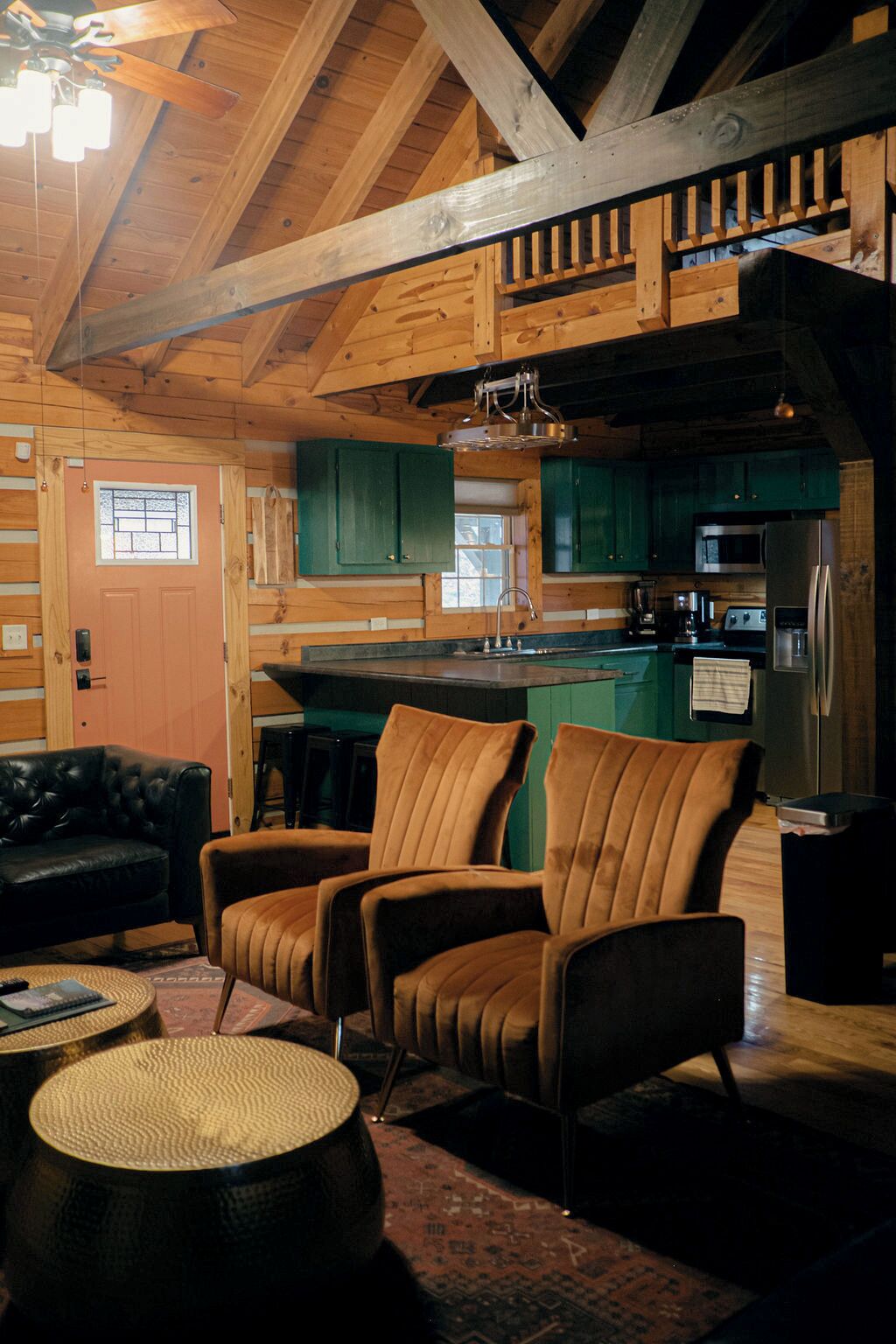 A cozy cabin interior with a wooden ceiling, a kitchen with green cabinets, and a seating area with leather chairs.