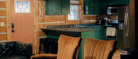 L’intérieur chaleureux d’un chalet avec un plafond en bois, une cuisine aux armoires vertes et un coin salon doté de fauteuils en cuir.