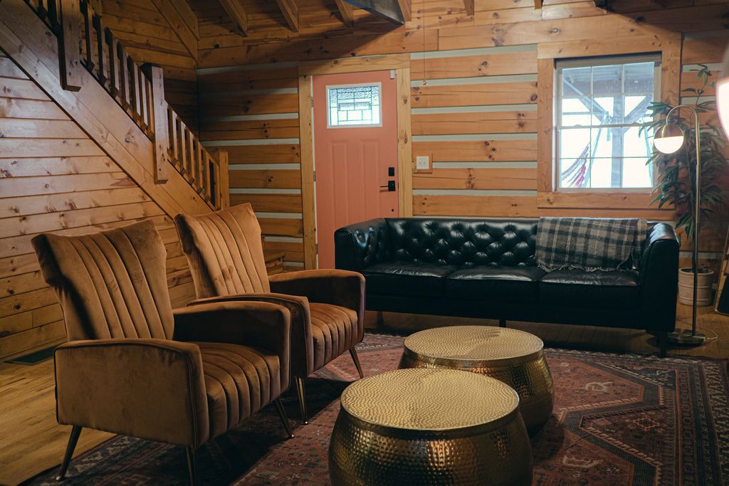 A log cabin interior with a staircase, a sofa, two armchairs, and a patterned rug.