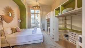 Ein Hotelzimmer mit Bett, Schreibtischlampe, Spiegel, einem Fenster mit Vorhängen und einem eingebauten Kleiderschrank mit Regalen.