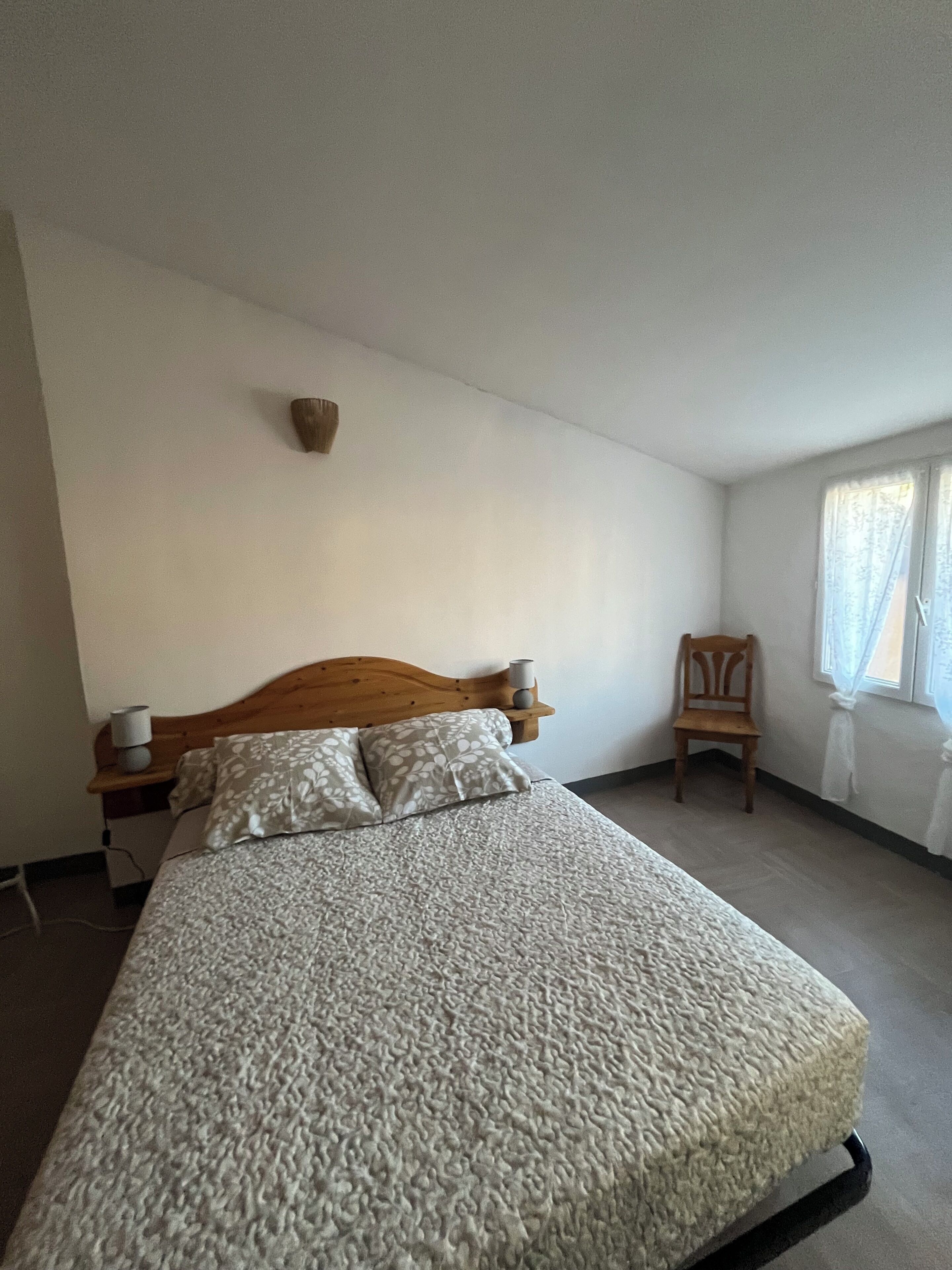 A bedroom with a bed, a wooden headboard, a bedside table with a lamp, a chair, and a window with curtains.