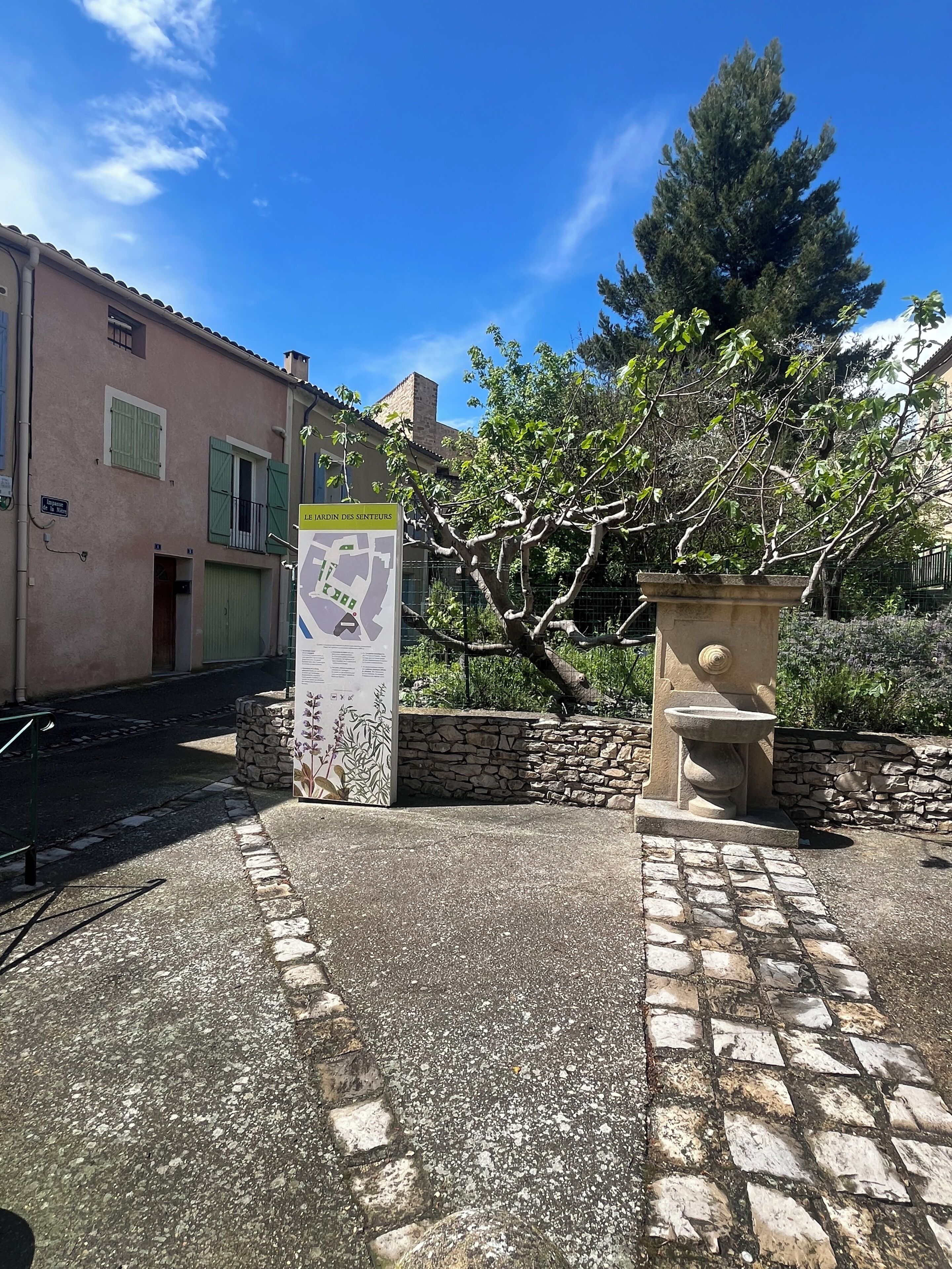 Une cour avec une fontaine en pierre, un panneau indicateur et un bâtiment aux volets verts.