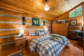 A cozy wooden cabin bedroom with a bed, nightstands, a dresser, and a window with a view of trees. - View  Hot Tub-Sauna-Fire Pit-Fireplace-Honeymoon (Sevierville)