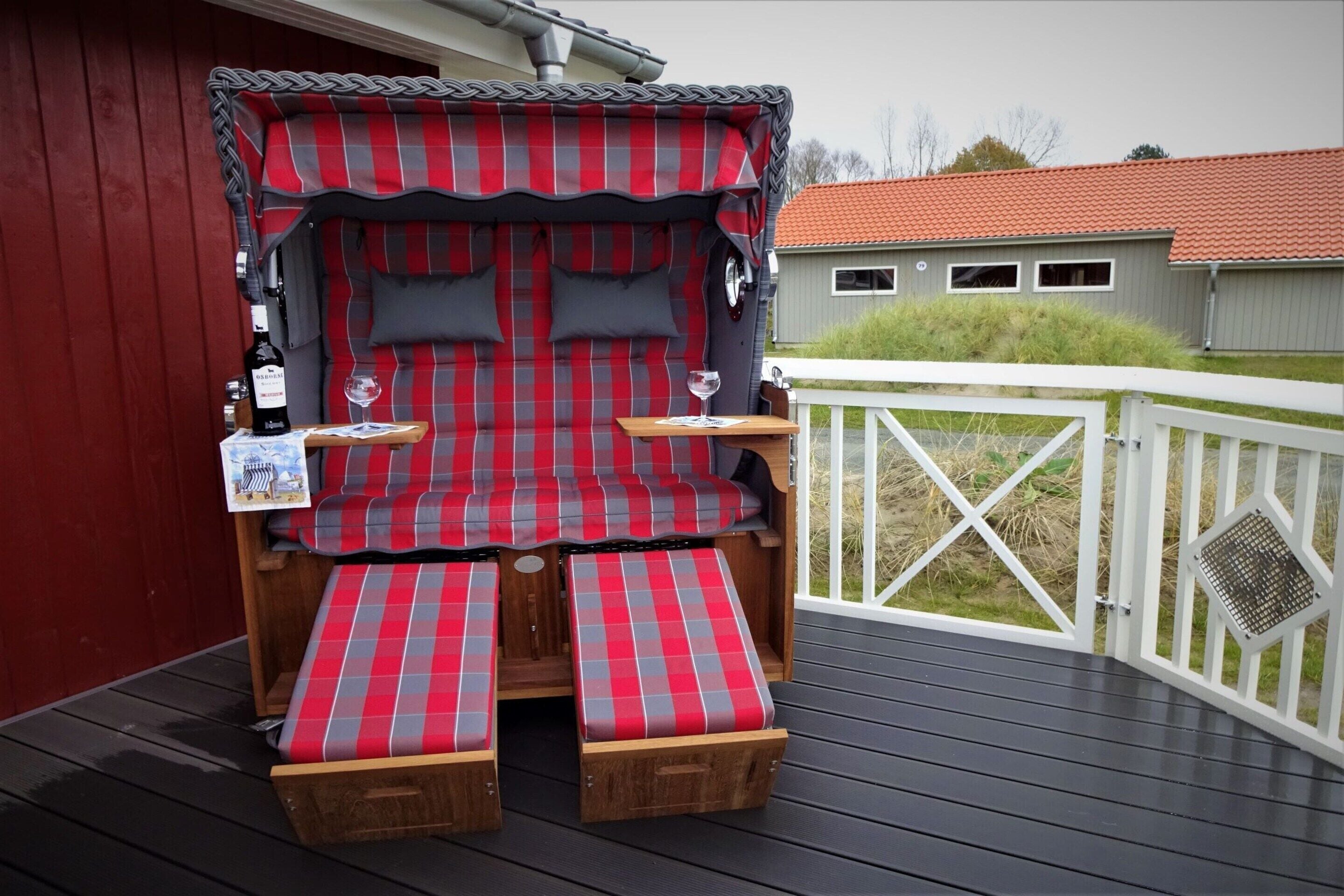 Un espacio al aire libre acogedor con un sofá rojo y gris de cuadros, dos bancos de madera y una mesa pequeña con una botella y vasos.