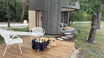 Une terrasse en bois avec une chaise blanche et une table dressée avec de la nourriture et des boissons, le tout entouré d’une zone boisée.