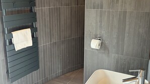 A modern bathroom with a dark tiled wall, a towel rack with a white towel, a toilet paper holder, and a white sink with a chrome faucet. - Oomi (Barbaste)