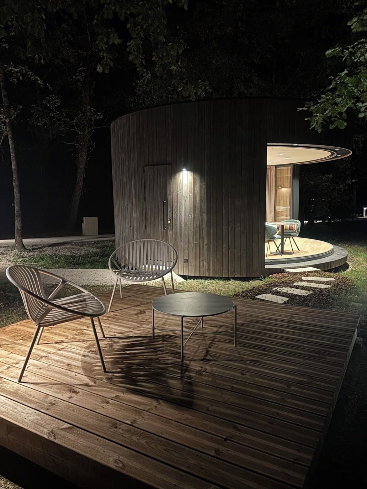 An outdoor wooden deck with modern furniture, a circular seating area, and a building with a door and illuminated interior.
