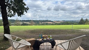 En picnicopstilling med et bord og stole på en træterrasse, med udsigt over en grøn mark og bakker.