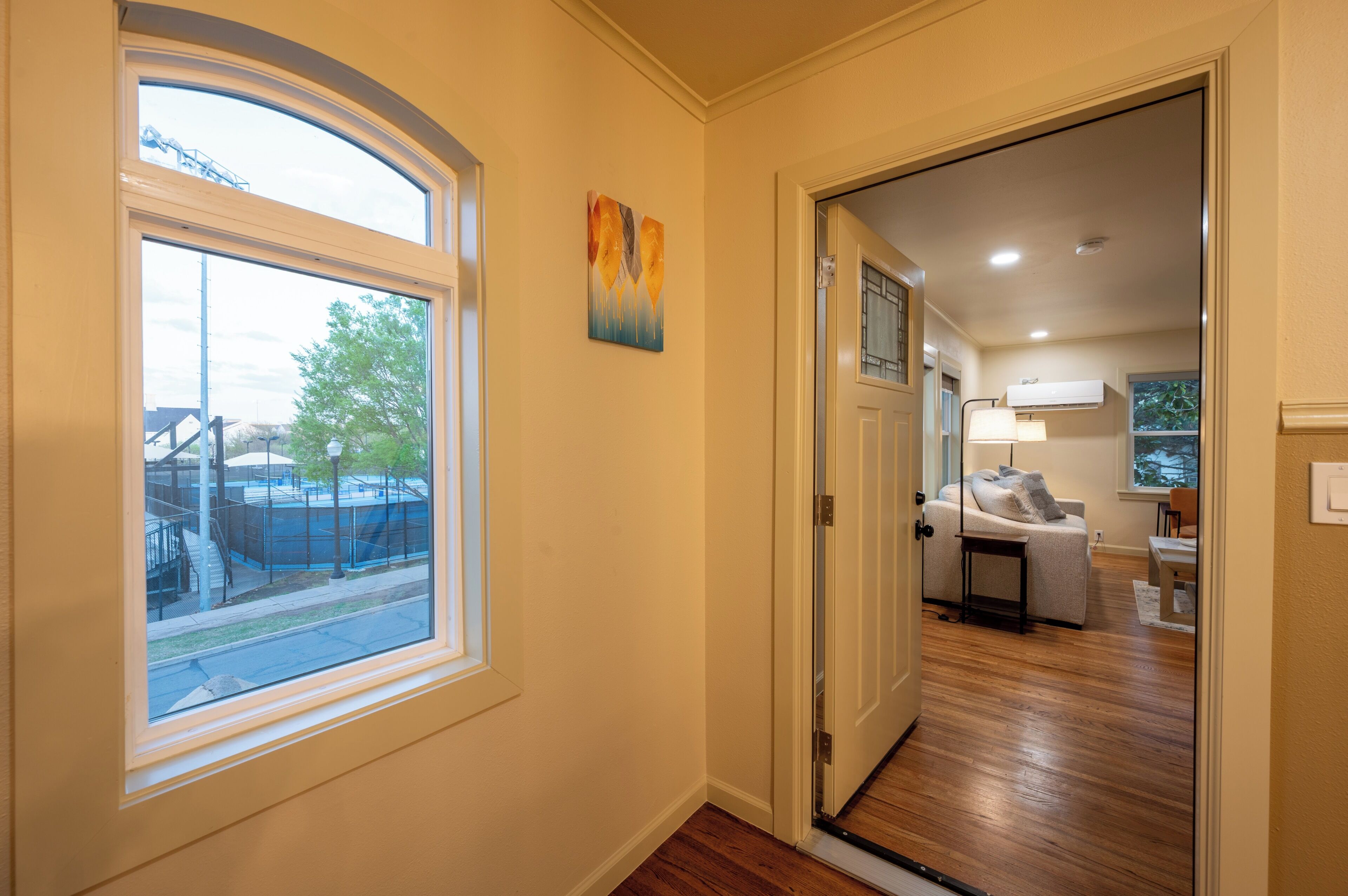 A hallway with a window, a door leading to a living area with a sofa and table, and a painting on the wall.