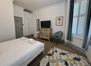Ein Hotelzimmer mit einem Bett, einem Schminktisch, einem Fernseher, einem Sessel und einem Fenster mit Jalousien.