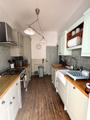 A kitchen with a stove, microwave, and wooden countertops. There is a clock on the wall and a shelf with dishes.