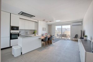Eine moderne Küche mit einer zentralen Kochinsel, einem Essbereich mit Stühlen und einer Glasschiebetür, die auf einen Außenbalkon führt.