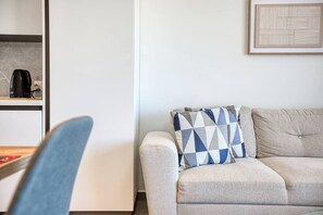 Ein modernes Wohnzimmer mit einer Couch, einem gemusterten Sitzkissen, einem gerahmten abstrakten Kunstwerk an der Wand und einer Kaffeemaschine auf einem Regal.