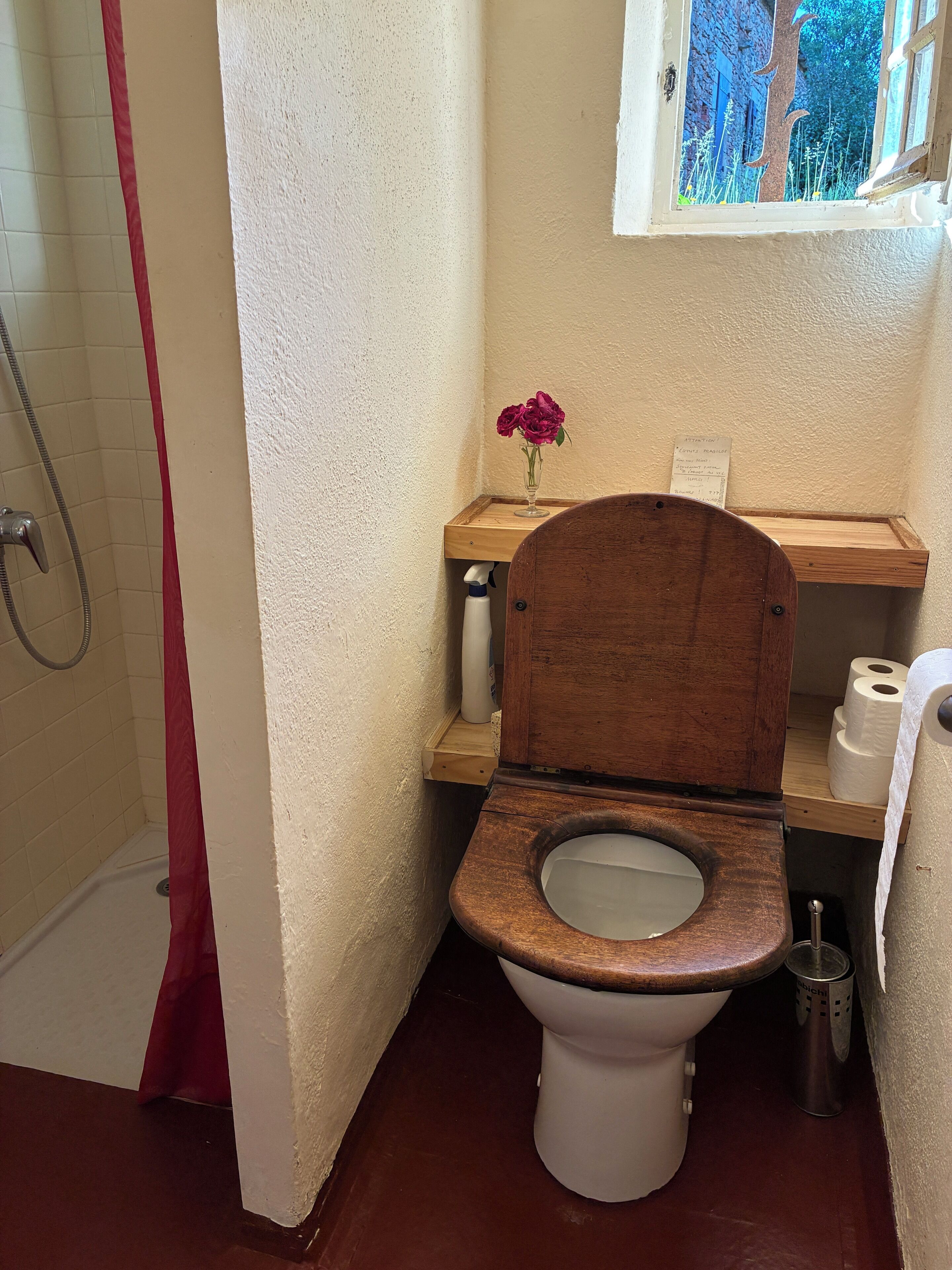 A bathroom with a wooden toilet, a shower, and a window with a view of trees.