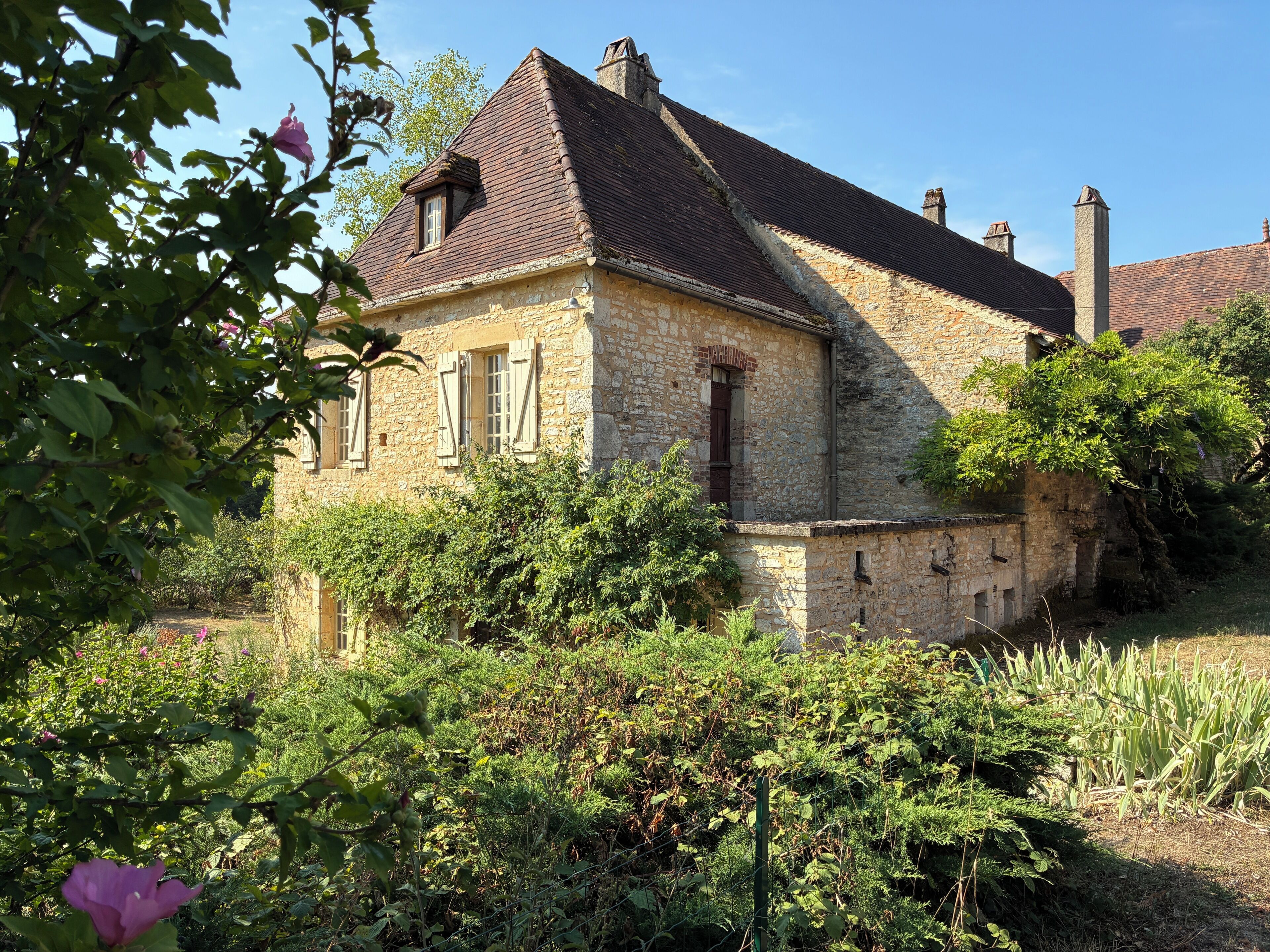 Un bâtiment en pierre doté d’un toit à deux versants, entouré d’une végétation luxuriante et d’un jardin.
