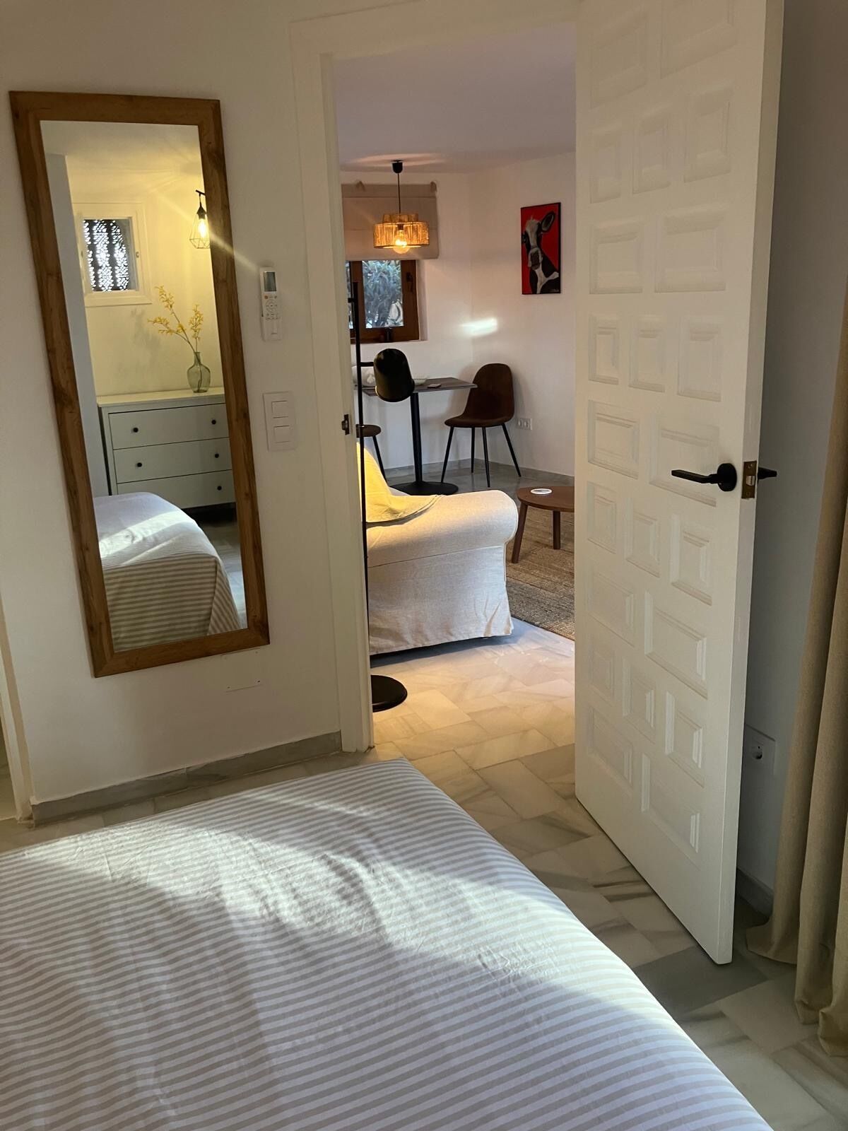 A bedroom with a large mirror, a bed with striped bedding, a small table with chairs, and a door leading to another room.