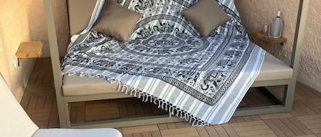 A sunlit outdoor seating area with a cushioned bench, a table, and a covered sleeping area with patterned bedding.