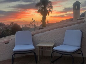 Zwei weiße Lounge-Stühle mit einem kleinen Tisch, vor einem Sonnenuntergang mit einer Palme und einem Gebäude.