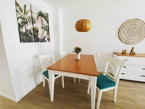 A dining area with a wooden table, white chairs, a hanging gold pendant light, and a wall with a tropical plant painting. - Ibizan-Style House in Benicasim · 3 minutes from the beach · Pool and parking (Benicàssim)