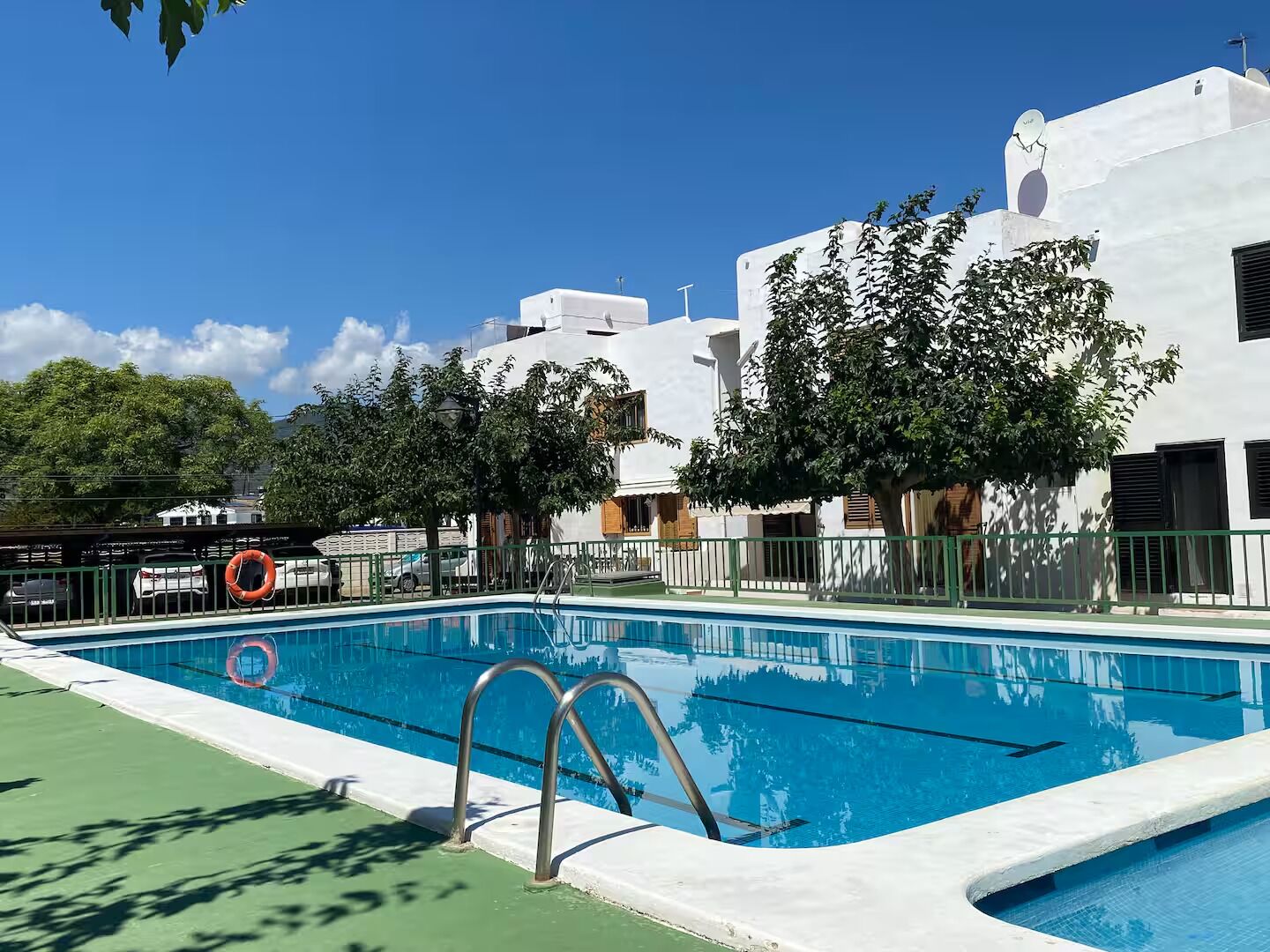 A swimming pool with a metal ladder, surrounded by a green fence, and adjacent to a white building with multiple windows and balconies.