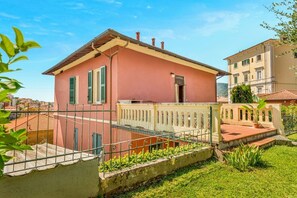 A pink villa with a balcony, surrounded by a white fence and greenery.