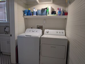 Een wasserette met twee witte voorladers, wasmachines en drogers, een plank met schoonmaakmiddelen en een raam met zonwering.