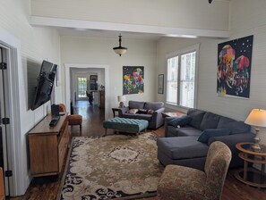 A spacious living room with a large sofa, a coffee table, and a television mounted on the wall. There is a patterned rug on the floor, and the room has a high ceiling with a hanging light fixture. - Historic 1908 Ybor City Bungalow (Tampa)