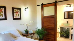 A bedroom with a bed, a nightstand, a potted plant, a sliding door, and a bathroom with a sink and mirror. - Casa Botanica (Lo de Marcos)