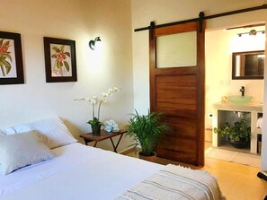 A bedroom with a bed, a nightstand, a potted plant, a sliding door, and a bathroom with a sink and mirror. - Casa Botanica (Lo de Marcos)