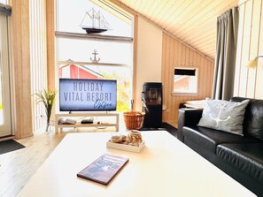 Ein gemütliches Wohnzimmer mit einem Flachbildfernseher, der eine Anzeige für einen Ferienort zeigt, einem Kamin, einem schwarzen Ledersofa und einem Couchtisch mit einem Buch und einem Korb.