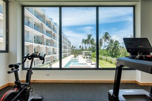 A room with a large window overlooking a pool and a building with balconies. There is a treadmill and an exercise bike inside. - Born | 2Bed Pool View | Up to 6 Guests (Bavaro)