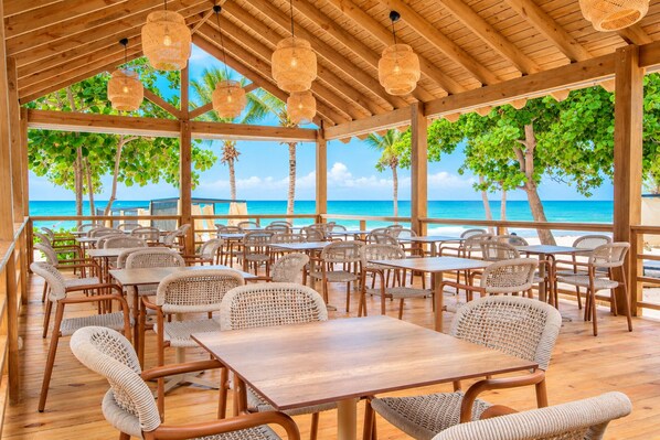 Ein überdachter Außenbereich mit Tischen und Stühlen, mit Blick auf einen Strand mit Palmen und das Meer.