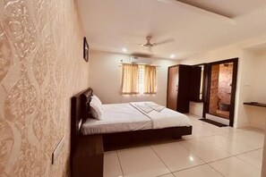 Un dormitorio con cama, ventilador de techo, ventana con cortinas y baño visible a través de una puerta abierta.