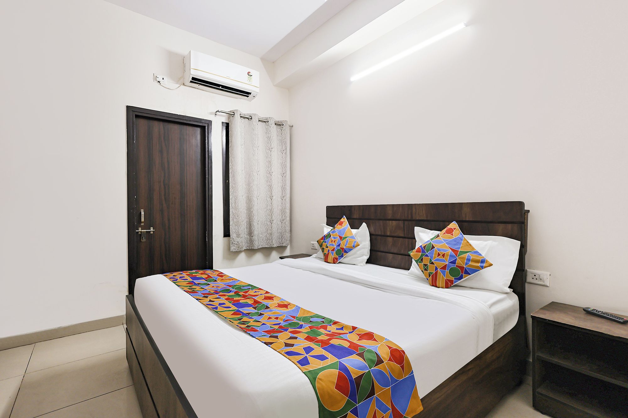Una habitación de hotel con una cama, una mesita de noche, un equipo de aire acondicionado, una puerta y una cortina.
