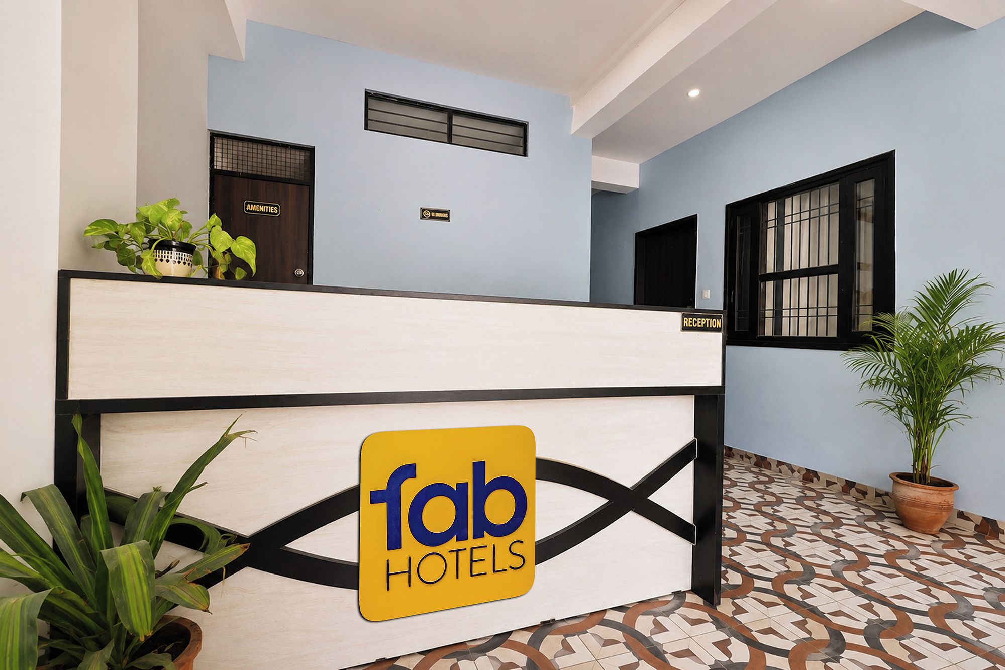 Un comptoir d’accueil d’hôtel avec une enseigne indiquant « fab hotels ». Des plantes en pot sont posées sur le comptoir, et une autre enseigne, plus petite, est fixée au-dessus.