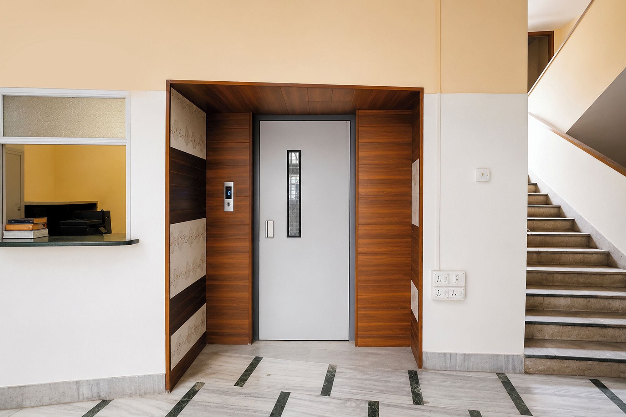 A modern interior with a wooden door, a digital lock, a staircase, and a wall-mounted shelf with books.