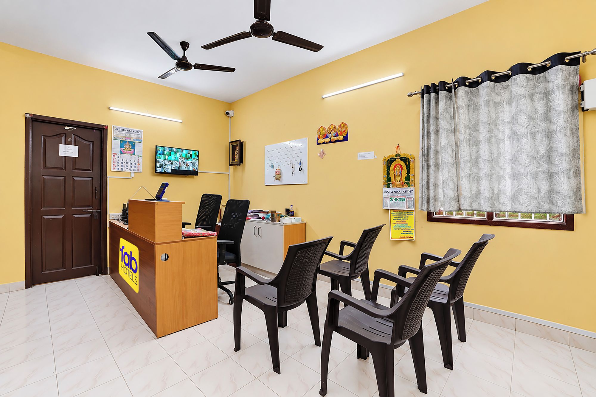A hotel reception area with a wooden desk, multiple chairs, ceiling fans, a television, and various wall decorations.