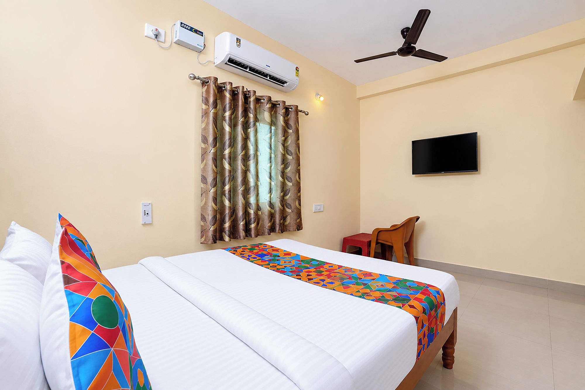 Une chambre d’hôtel équipée d’un lit, d’une télévision, d’un climatiseur, d’un ventilateur de plafond et d’une fenêtre avec des rideaux.