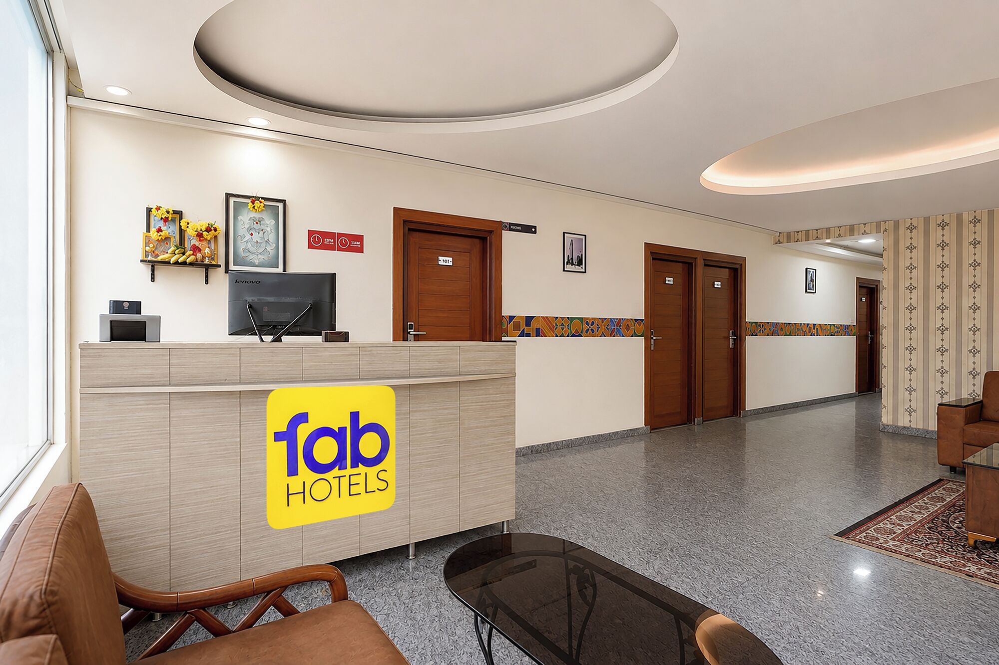 A hotel reception area with a reception desk, a seating area with chairs, and a corridor leading to doors.