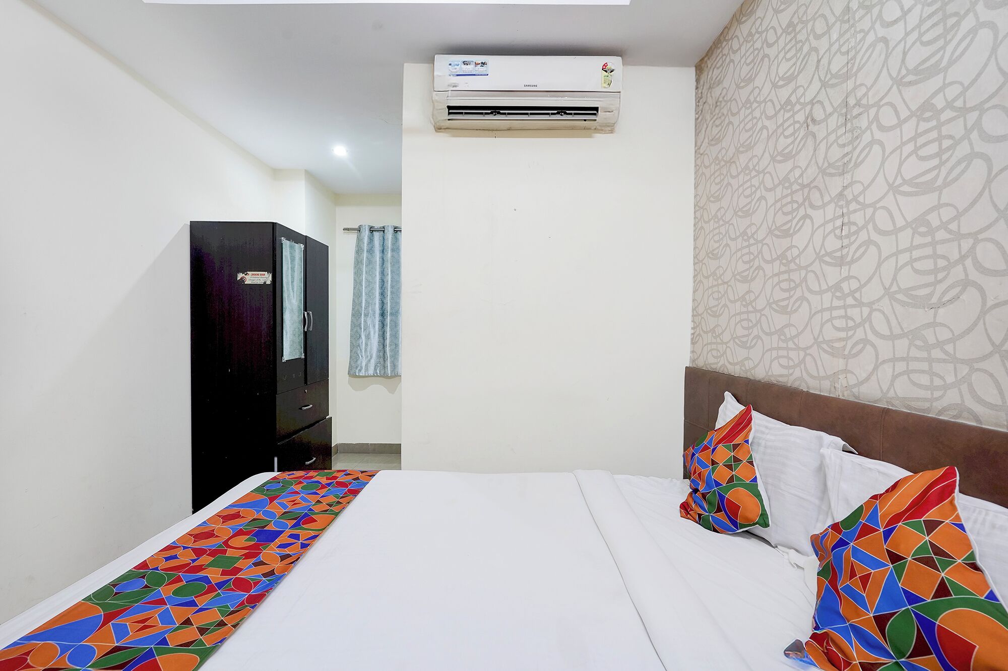 A hotel room with a bed, a wardrobe, an air conditioning unit, and a patterned wall.